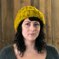 A person with shoulder-length dark hair is wearing the Monolith Hat by Halcyon Yarn, a chunky knit hat in mustard-yellow made from bulky weight yarn. They are looking directly at the camera with a neutral expression, standing against a backdrop of wooden panels.