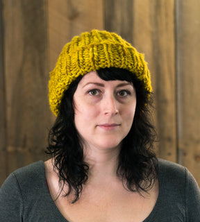 A person with shoulder-length dark hair is wearing the Monolith Hat by Halcyon Yarn, a chunky knit hat in mustard-yellow made from bulky weight yarn. They are looking directly at the camera with a neutral expression, standing against a backdrop of wooden panels.