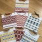 Seven intricately woven Halcyon Yarn baby bibs lay arranged on a wooden surface. The bibs feature various patterns in red, yellow, blue, green, and white with different geometric designs. Each bib has a braided or twisted tie for securing around the neck—a perfect stash buster project for any weaver utilizing the Woven Baby Bib Pattern download.