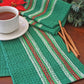 A white cup filled with tea sits on a green, red, and white striped cloth atop a wooden surface. Beside the cup, a folded matching Waffle Weave Dish Towel from Halcyon Yarn and a couple of cinnamon sticks are neatly placed. In the background, festive greenery and red poinsettias enhance the holiday theme.