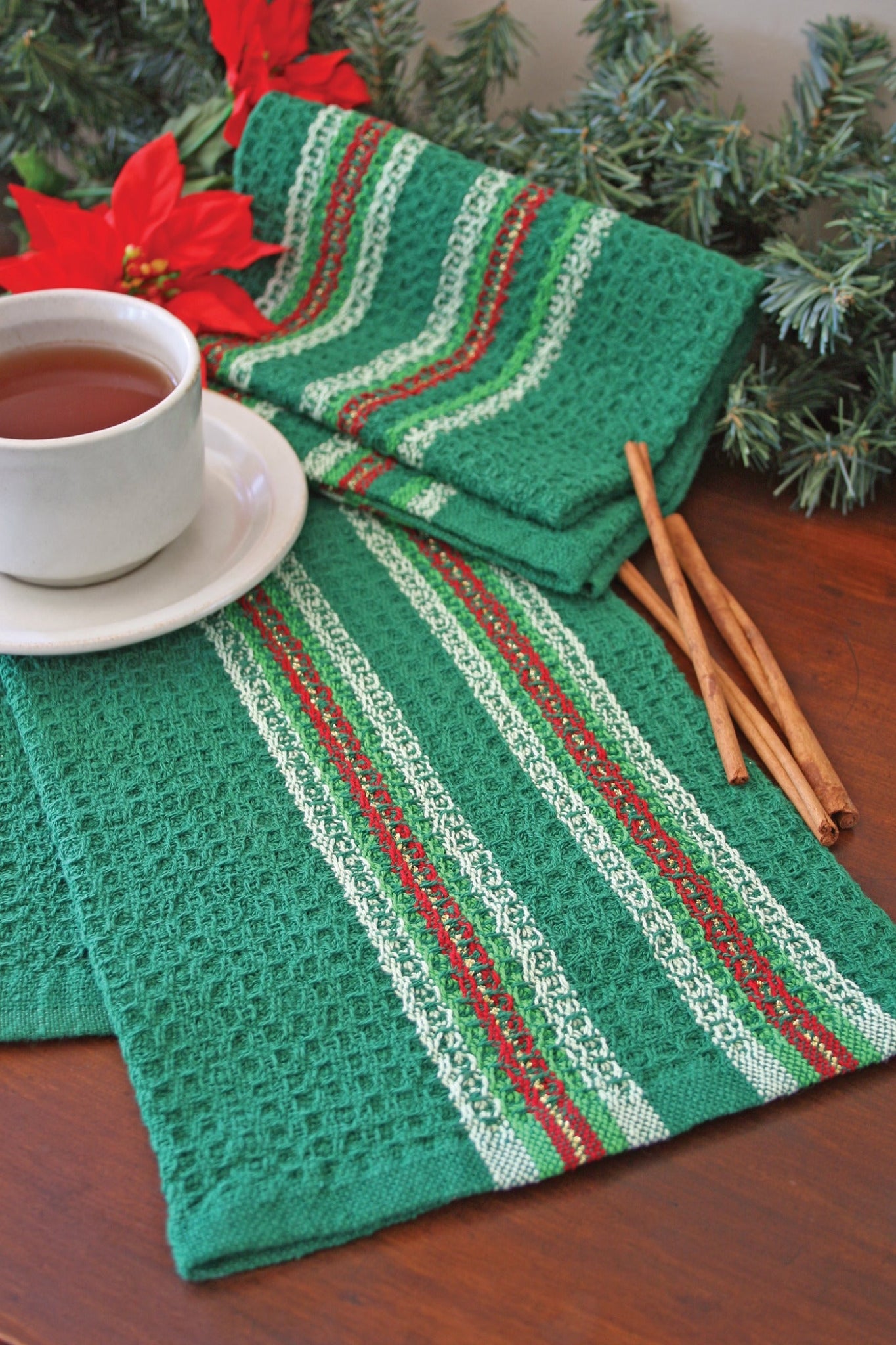 A white cup filled with tea sits on a green, red, and white striped cloth atop a wooden surface. Beside the cup, a folded matching Waffle Weave Dish Towel from Halcyon Yarn and a couple of cinnamon sticks are neatly placed. In the background, festive greenery and red poinsettias enhance the holiday theme.