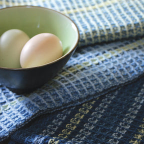 A small, green bowl containing one cream-colored egg and one light beige egg is placed on top of a stack of textured blue and green striped fabrics, featuring the waffle weave design from Halcyon Yarn's Waffle Weave Dish Towels kit.