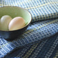 A small green bowl with two eggs sits on top of layered Waffle Weave Dish Towels by Halcyon Yarn. The towels, crafted with a four-shaft loom, feature different shades of blue and white stripes. The scene is set on a wooden surface.