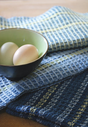 A small green bowl with two eggs sits on top of layered Waffle Weave Dish Towels by Halcyon Yarn. The towels, crafted with a four-shaft loom, feature different shades of blue and white stripes. The scene is set on a wooden surface.