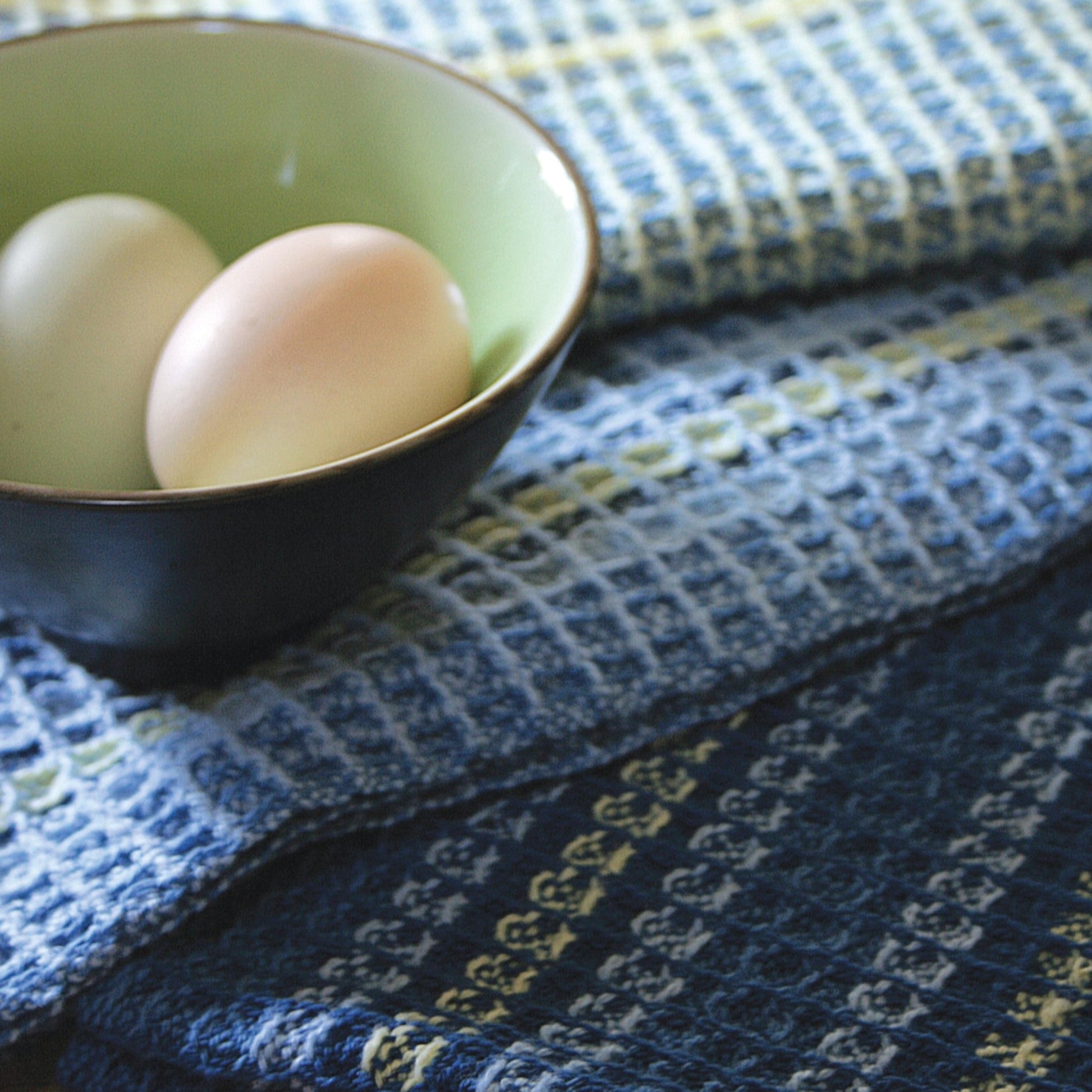 A small bowl containing two eggs is placed on Halcyon Yarn's Waffle Weave Dish Towels. The fabrics have a blue and white checkered pattern, and the bowl is green inside with a dark exterior, creating a cozy and homely ambiance.