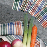 A collection of vegetables, including a red onion, a white turnip, two leeks, and a carrot, arranged on a plaid Homestead Cotton cloth. A knife sits beside the vegetables. Three additional folded Camp & Cottage Towels by Halcyon Yarn are placed in the background on a gray surface.