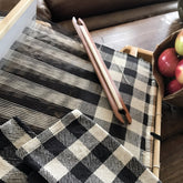 A close-up of black and white checkered fabric woven on a 4-shaft loom, with a shuttle resting atop. To the right, a paper bag filled with red apples partially visible on a wooden table evokes the charm of a cozy kitchen, where one might find Halcyon Yarn's Buffalo Plaid Woven Dish Towels in progress.