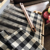 A close-up of a woven Buffalo Plaid Woven Dish Towel from Halcyon Yarn in black and white being worked on a four-shaft loom. A wooden shuttle used for weaving is resting on the fabric. In the background, there is a bowl containing red apples.