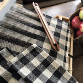 A traditional four-shaft loom from Halcyon Yarn is in use, weaving a fabric that closely resembles the black and white checkered pattern of the Buffalo Plaid Dish Towel Kit. Wooden tools lie on the loom, while a paper bag filled with fresh apples sits nearby on a wooden surface.