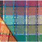 The Homestead Cotton Color Gamp from Halcyon Yarn is a vibrant woven textile showcasing a complex, multicolored plaid pattern in shades of red, orange, yellow, green, blue, and purple. This fabric could be ideal for creating a lively set of kitchen towels. It is slightly folded at the bottom left corner, demonstrating the continuity of the pattern across the fold.