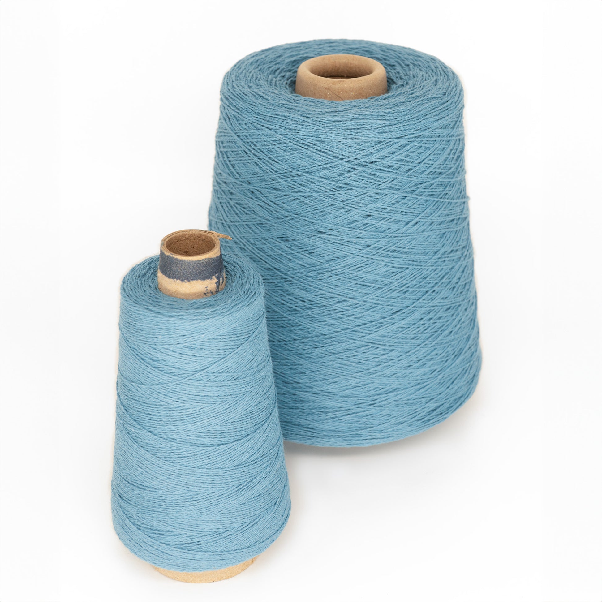 Two spools of light blue, machine-washable Homestead 8/2 Cotton Yarn from Supreme Corp are shown against a plain white background. The spool on the left is smaller and slightly shorter than the larger cone on the right. Both spools have tan-colored cores.