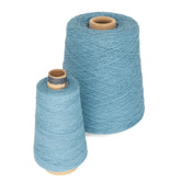 The scene features two spools of light blue cotton yarn from Supreme Corp against a white background. The Homestead 8/2 Cotton Yarn on the large cone, perfect for weaving projects, stands prominently behind the smaller spool. Both display cardboard cores, highlighting the texture of the Homestead Cotton yarn.