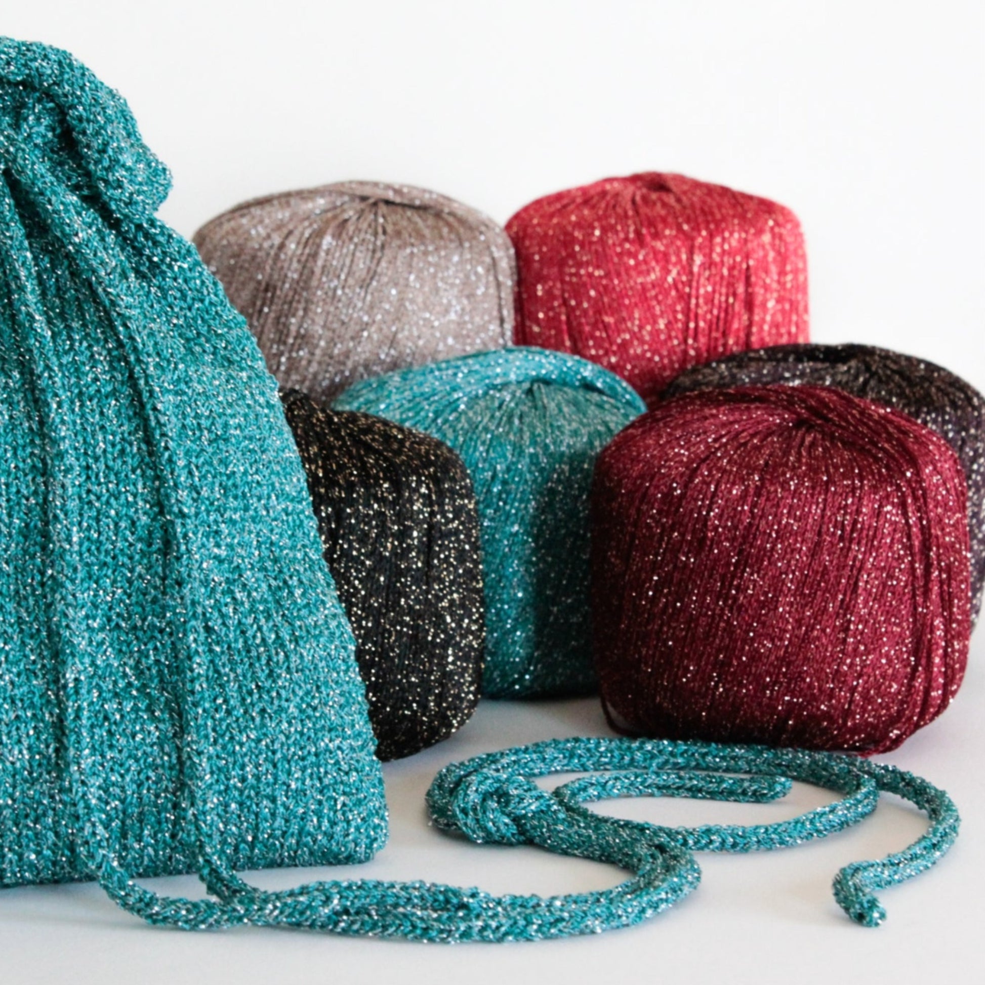 Various skeins of sparkly yarn in colors including teal, silver, red, black, pink, and burgundy rest together on a white surface. In the foreground, the A Bit of Sparkle Evening Bag from Halcyon Yarn gleams in teal—ideal for holding your night-out essentials or gifting as a quick-to-knit present.