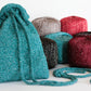 The "A Bit of Sparkle Evening Bag" by Halcyon Yarn, crafted from teal, sparkly knit fabric, is prominently displayed in the foreground. Behind the bag are six balls of yarn in diverse colors—teal, beige, burgundy, black, and red—all featuring a subtle shimmer. Ideal for a quick-to-knit gift or carrying night-out essentials. The scene is set against a plain white backdrop.