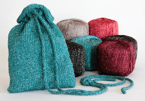 The "A Bit of Sparkle Evening Bag" by Halcyon Yarn, crafted from teal, sparkly knit fabric, is prominently displayed in the foreground. Behind the bag are six balls of yarn in diverse colors—teal, beige, burgundy, black, and red—all featuring a subtle shimmer. Ideal for a quick-to-knit gift or carrying night-out essentials. The scene is set against a plain white backdrop.