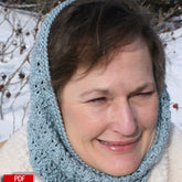 A woman with short brown hair smiles while wearing the "Fountain Hood" by Halcyon Yarn, a light blue knitted headscarf against a snowy background. The cozy headscarf, featuring a detailed lace pattern, doubles as a cowl hood. Snow-covered bushes with small red berries are visible in the background.