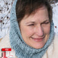 A woman wearing the Fountain Hood in light blue, designed by Halcyon Yarn, smiles outdoors in a snowy landscape. She is dressed in a warm beige coat with a fur collar, exuding a luxurious and cozy vibe. A "download" icon along with an Adobe PDF logo appear at the bottom left corner of the image.