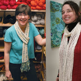 Two women are pictured wearing the elegant Silk & Sari Ribbon Scarf from Halcyon Yarn. The woman on the left, in a blue shirt and glasses, stands in front of shelves filled with yarn. The woman on the right, in a red sweater, stands near a wall displaying artwork with a floral design.