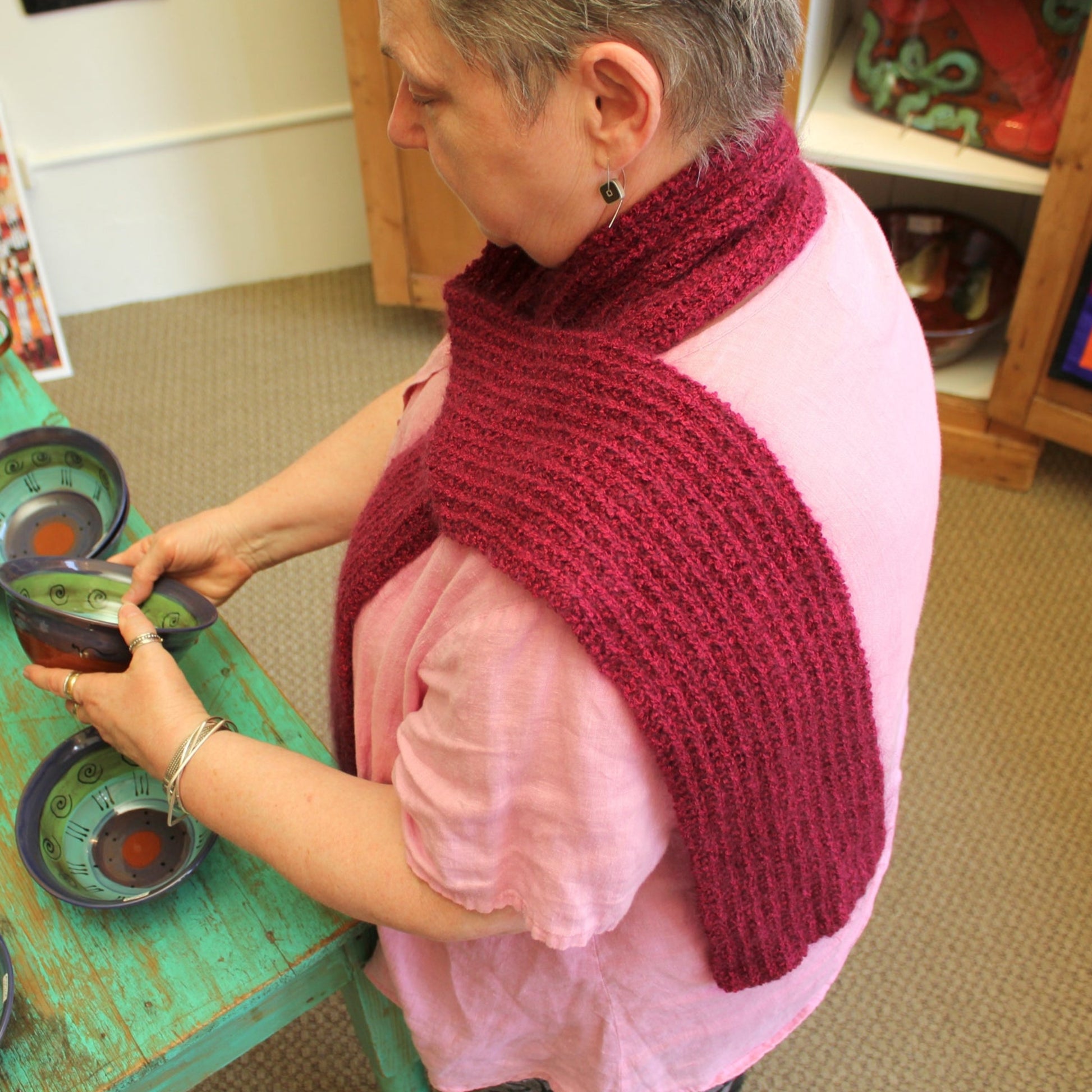 A person with short hair, clad in a pink shirt and sporting a Reversible Silk Bouclé Scarf from Halcyon Yarn around their neck, is examining canned goods placed on a turquoise table in a room filled with shelves stocked with various items. The cozy and casual atmosphere is enhanced by the stylish reversible scarf they're wearing.