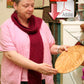 A person wearing a pink shirt and a Halcyon Yarn Reversible Silk Bouclé Scarf holds a wooden plate in a shop. Various items, including textiles and ceramics, are displayed around them. An Adobe PDF download icon is digitally overlaid on the image.