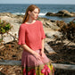 A woman with long hair sits on a wooden log near a rocky beach. She is wearing a coral-colored Petal Sleeve Top from Halcyon Yarn and a Signature Gemstone Silk Boucle skirt with horizontal stripes and floral designs. The background showcases the ocean, rocks, and some dried shrubs, capturing the essence of silky comfort.