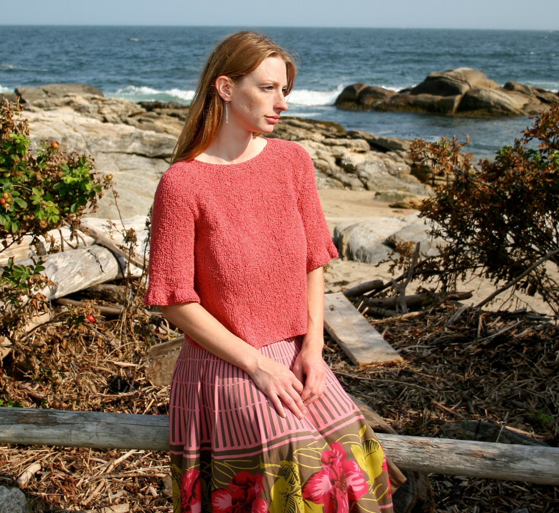 A woman with long hair sits on a wooden log near a rocky beach. She is wearing a coral-colored Petal Sleeve Top from Halcyon Yarn and a Signature Gemstone Silk Boucle skirt with horizontal stripes and floral designs. The background showcases the ocean, rocks, and some dried shrubs, capturing the essence of silky comfort.