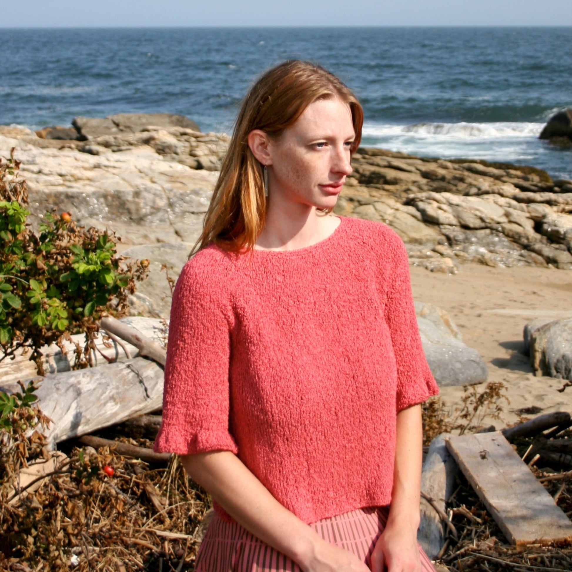 A person with long hair, wearing the Halcyon Yarn Petal Sleeve Top in bright coral silk bouclé paired with a striped skirt, stands near a rocky beach with the ocean in the background. The clear sky suggests a sunny day, and the person gazes off to the side with a serene expression.