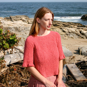 A woman with shoulder-length hair stands on a rocky beach with the ocean in the background. She is wearing a Halcyon Yarn Petal Sleeve Top in Silk Bouclé and a pink pleated skirt with feminine ruffles. She looks thoughtful as she gazes into the distance.