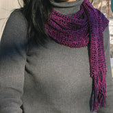 A person wearing a dark turtleneck sweater and a luxurious, textured purple Lacy Leno Silk Scarf from Halcyon Yarn with fringes stands in an indoor setting. Only the torso, shoulders, and part of the head are visible, with the face not shown. The background hints at a bright, possibly sunlit area.