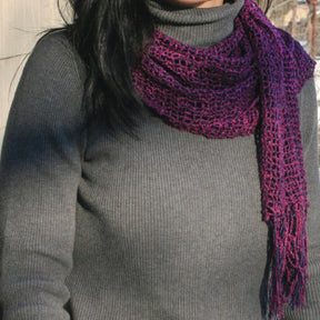 A person wearing a grey turtleneck sweater is draped in the luxurious Halcyon Yarn Lacy Leno Silk Scarf. The magenta scarf, adorned with a leno lace pattern and finished with fringed ends, hangs loosely around their neck. The person's face is not visible in the image.