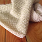 A close-up of a knitted blanket in a soft, light beige color draped over a wooden table showcases the Royal Quilt Stitch pattern. The blanket, reflecting the design of Halcyon Yarn's Royal Quilt Stitch Cowl, has a textured honeycomb design. Beside it is a skein of matching yarn. The table surface features a natural wood grain.