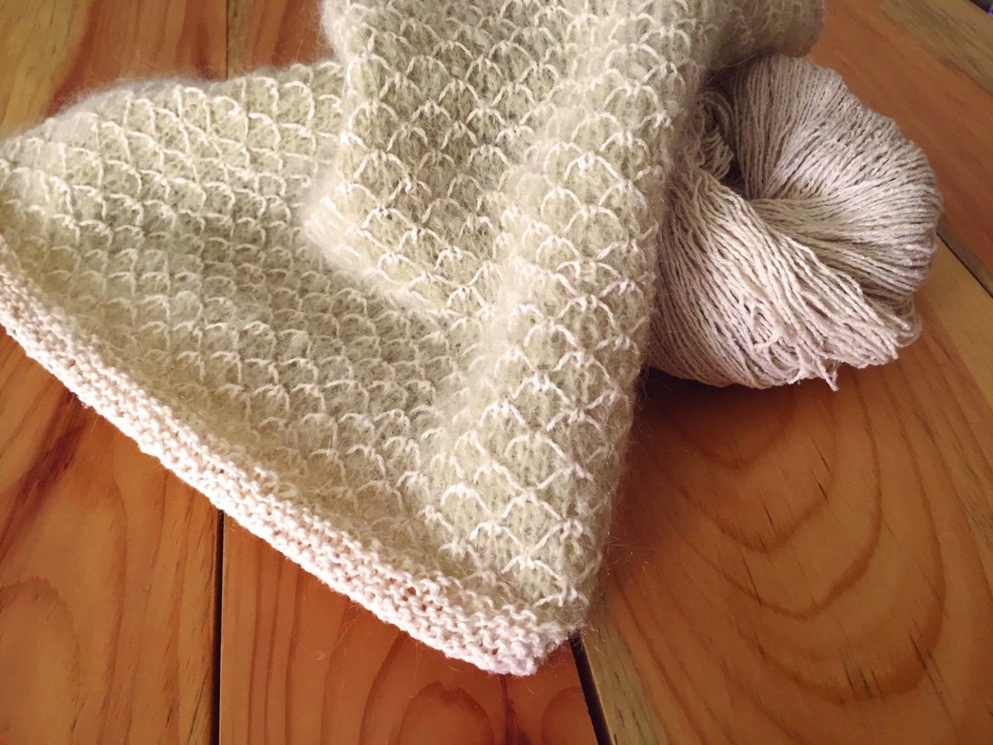 A close-up of a knitted blanket in a soft, light beige color draped over a wooden table showcases the Royal Quilt Stitch pattern. The blanket, reflecting the design of Halcyon Yarn's Royal Quilt Stitch Cowl, has a textured honeycomb design. Beside it is a skein of matching yarn. The table surface features a natural wood grain.