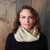 A person with wavy, shoulder-length hair is facing the camera with a slight smile. They are wearing a cream-colored Royal Quilt Stitch Cowl from Halcyon Yarn over a dark top and are posed against a rustic wooden background.