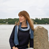 A woman with long brown hair wearing a blue sweater and a Silky Honeycomb Scarf by Halcyon Yarn sits on a rock by a calm body of water, with trees lining the opposite shore under a cloudy sky.