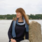 A person with long hair, wearing a dark sweater and the luxurious Silky Honeycomb Scarf by Halcyon Yarn in blue, sits on a rock by a calm body of water. In the background, trees and a cloudy sky frame the scene as the individual gazes to the side, away from the camera.