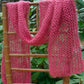 The Silky Honeycomb Scarf by Halcyon Yarn, a delicate pink and luxurious accessory, is draped over the back of a wooden chair with a woven seat. The scarf features an elegant lacy, open-knit pattern and is displayed against a backdrop of green foliage.