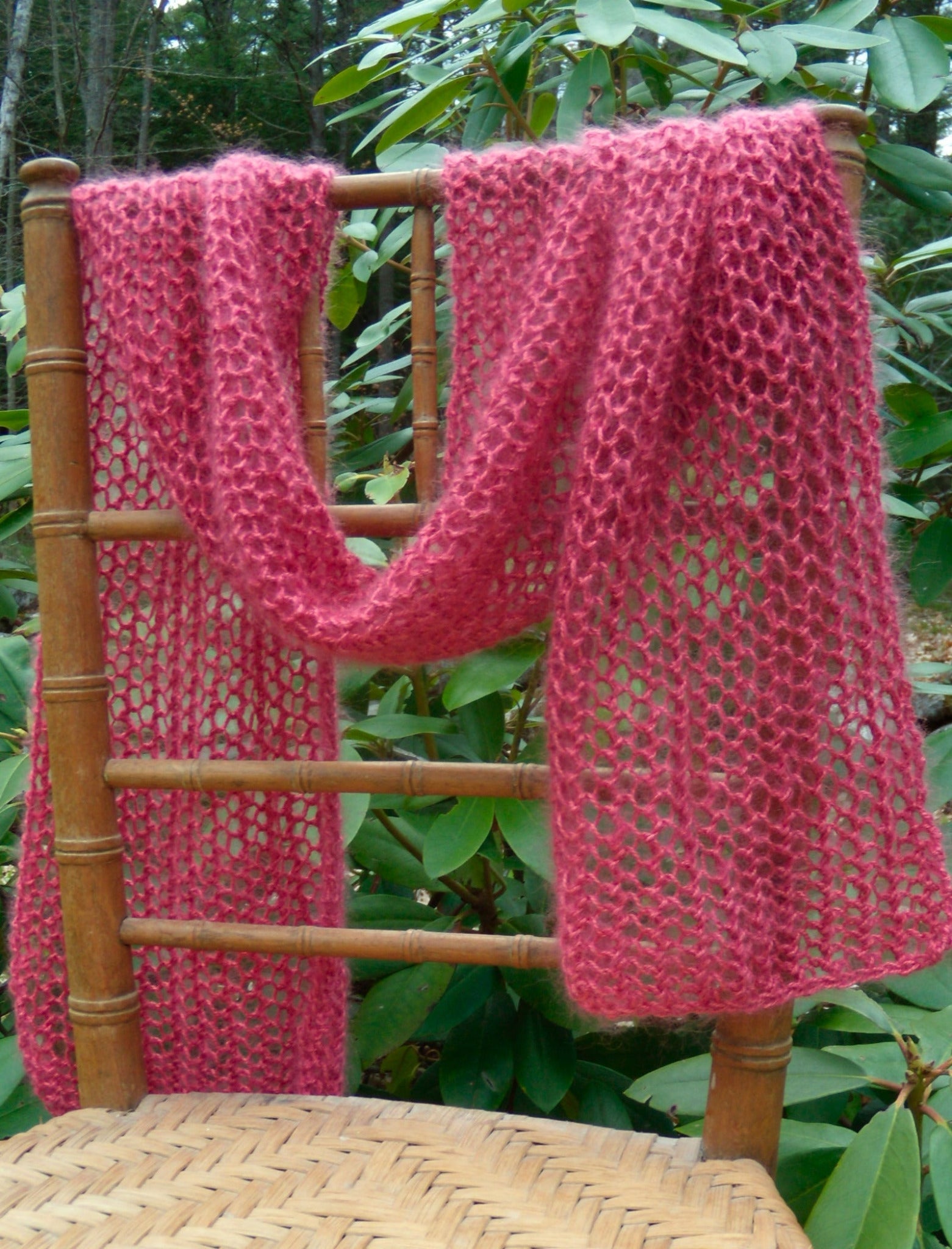 The Silky Honeycomb Scarf Pattern by Halcyon Yarn, a luxurious accessory in a soft pink hue, is gracefully draped over the back of a wooden chair. Nestled within a garden of lush green foliage, it exhibits exquisite color combinations that enhance its charm.