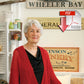 A senior woman with short gray hair, dressed in a red Halcyon Yarn Honeycomb Kimono that appears to be a mohair/silk blend and a black top, stands indoors before a wall decorated with vintage signs. A large Adobe PDF icon featuring a red arrow is overlaid in the top-right corner of the image.