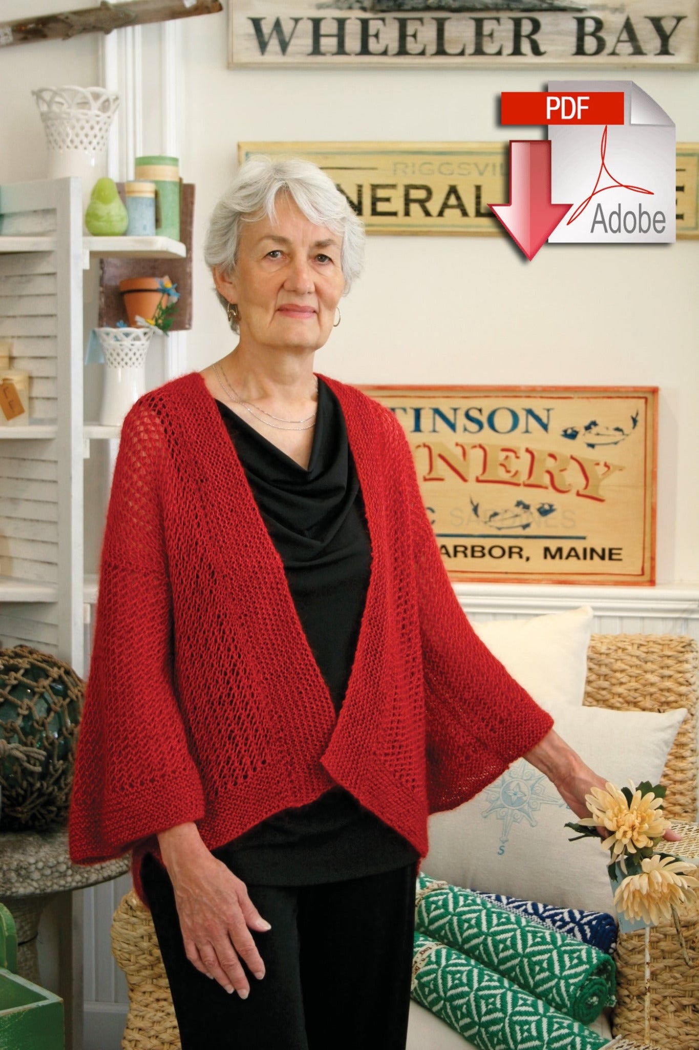 An elderly woman with short gray hair is wearing a Halcyon Yarn Honeycomb Kimono, made from a cozy mohair/silk blend, over a black top. She stands in a room decorated with various signs and coastal-themed items. In the upper right corner of the image, there is an Adobe PDF logo with a red arrow pointing down, indicating the option to download.