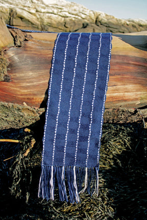 The Seguin Sparkling Waves Scarf by Halcyon Yarn, an elegant woven piece in dark blue with light blue and white stripes, is draped over a large log. The scarf, featuring fringes at the ends, is displayed outdoors against the Rocky terrain and sparse vegetation reminiscent of Seguin Island Light.