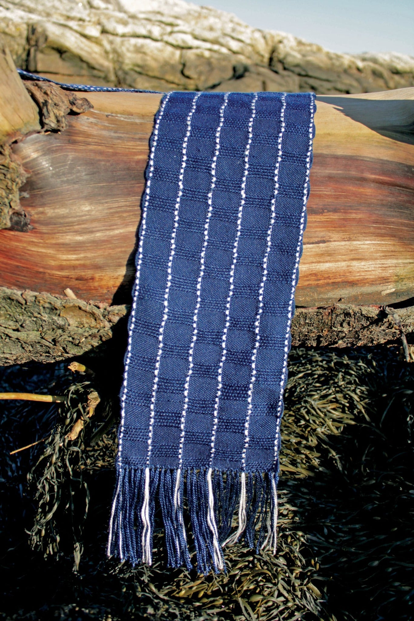 The Seguin Sparkling Waves Scarf by Halcyon Yarn, a dark blue woven silk accessory adorned with white stripes and fringed ends, is elegantly draped over a large piece of weathered driftwood. In the background, rugged rocks covered in patches of snow stand against a clear sky reminiscent of Seguin Island Light. The foreground features dark seaweed scattered across the ground.