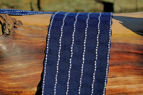 A close-up of the Seguin Sparkling Waves Scarf by Halcyon Yarn resting on a wooden log. The fabric is dark blue with vertical rows of white dashed lines, creating a striped pattern. The log has a smooth, polished surface with natural wood grain and varying shades of brown.