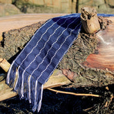 A Seguin Sparkling Waves Scarf by Halcyon Yarn, in an elegant navy blue with white stripes, lies draped over a piece of weathered driftwood. The scarf features fringe tassels at the ends and is displayed under bright sunlight, highlighting its texture against the natural, rustic background near Seguin Island Light.