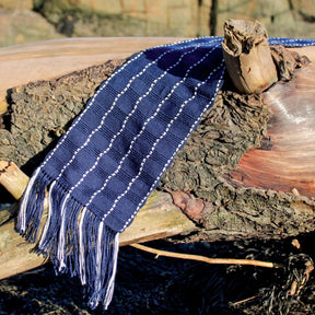 A Seguin Sparkling Waves Scarf by Halcyon Yarn, in an elegant navy blue with white stripes, lies draped over a piece of weathered driftwood. The scarf features fringe tassels at the ends and is displayed under bright sunlight, highlighting its texture against the natural, rustic background near Seguin Island Light.