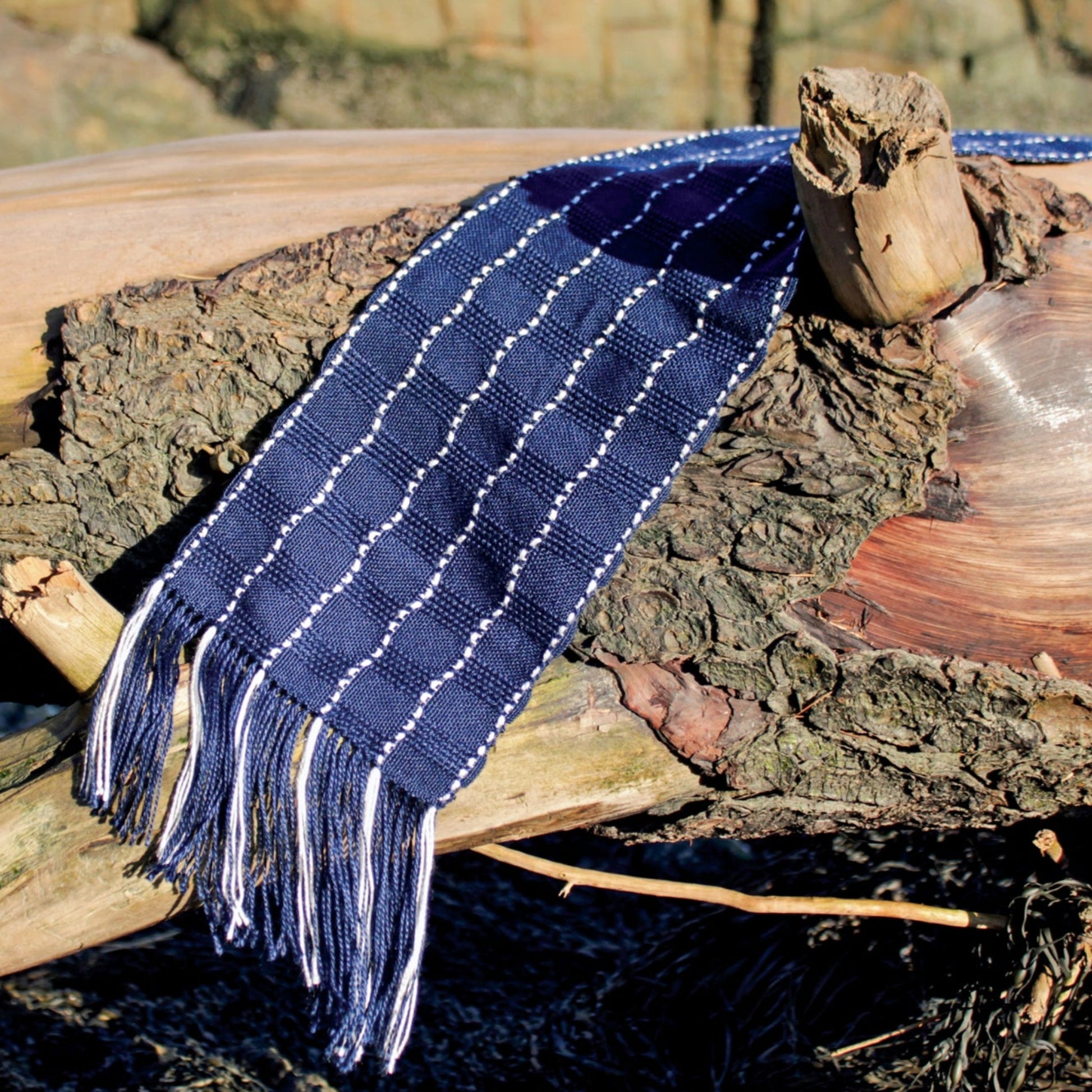 A Seguin Sparkling Waves Scarf from Halcyon Yarn, featuring dark blue woven silk with white checkered patterns and fringed ends, is draped over a weathered log. The texture of the scarf sharply contrasts with the rough, bark-covered wood, while some rocks appear blurred in the background. Sunlight highlights the elegant details of this accessory near Seguin Island Light.