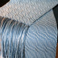 A close-up view of a textured, handwoven Snow Crystal Shadow Weave Scarf by Halcyon Yarn draped over a surface. The scarf features a light blue and white zigzag pattern with fringed ends. The shimmering shadow weave adds depth, while the intricate design and soft colors give the scarf a cozy and elegant appearance.