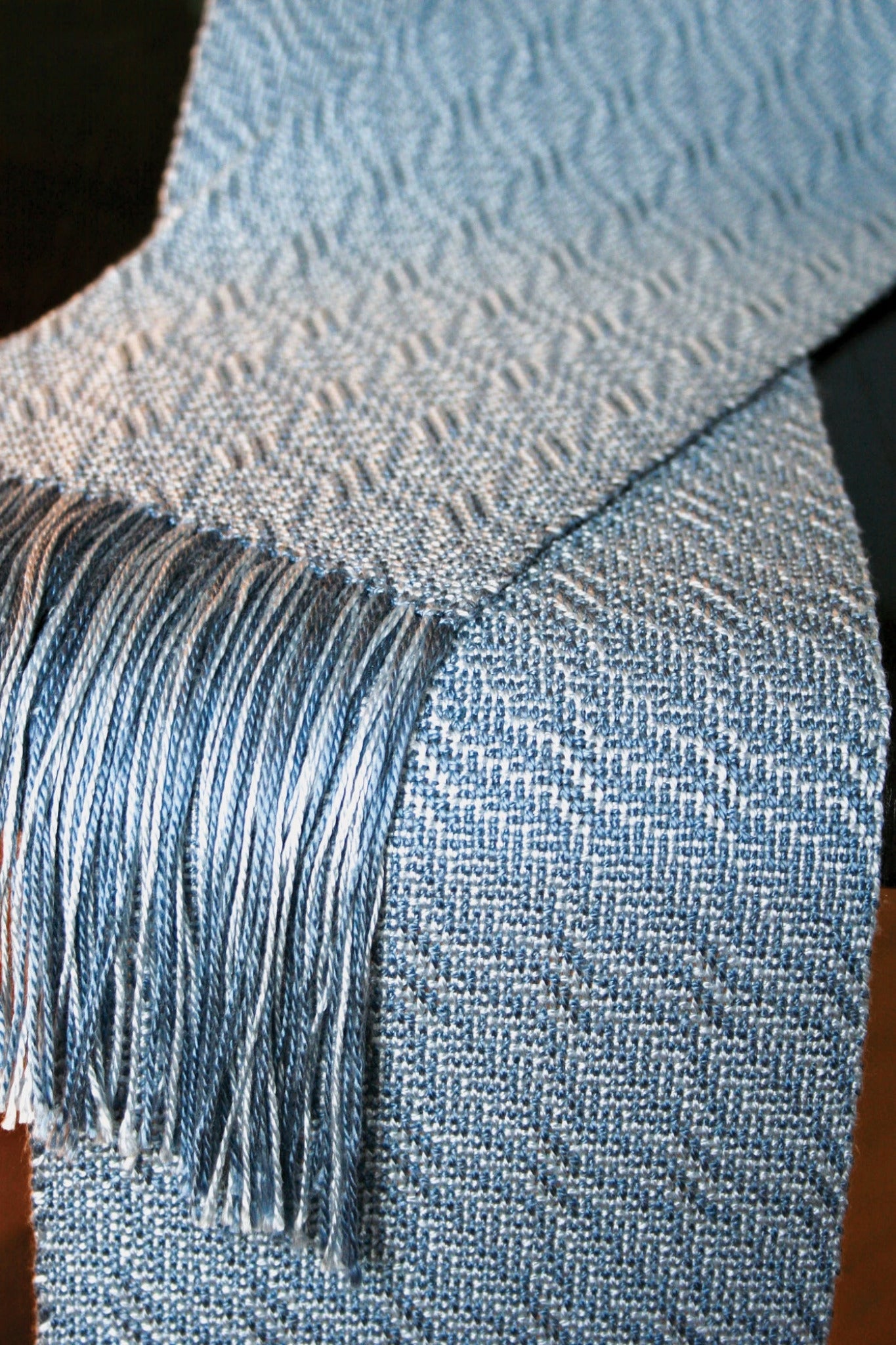 A close-up view of a textured, handwoven Snow Crystal Shadow Weave Scarf by Halcyon Yarn draped over a surface. The scarf features a light blue and white zigzag pattern with fringed ends. The shimmering shadow weave adds depth, while the intricate design and soft colors give the scarf a cozy and elegant appearance.