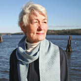 A woman with short white hair and a Snow Crystal Shadow Weave Scarf from Halcyon Yarn stands in front of a body of water, gazing into the distance. She is wearing a dark top that seems to shimmer like 2/12 Gemstone Silk. The background shows a hilly shoreline with wooden pylons, all beneath a clear blue sky.