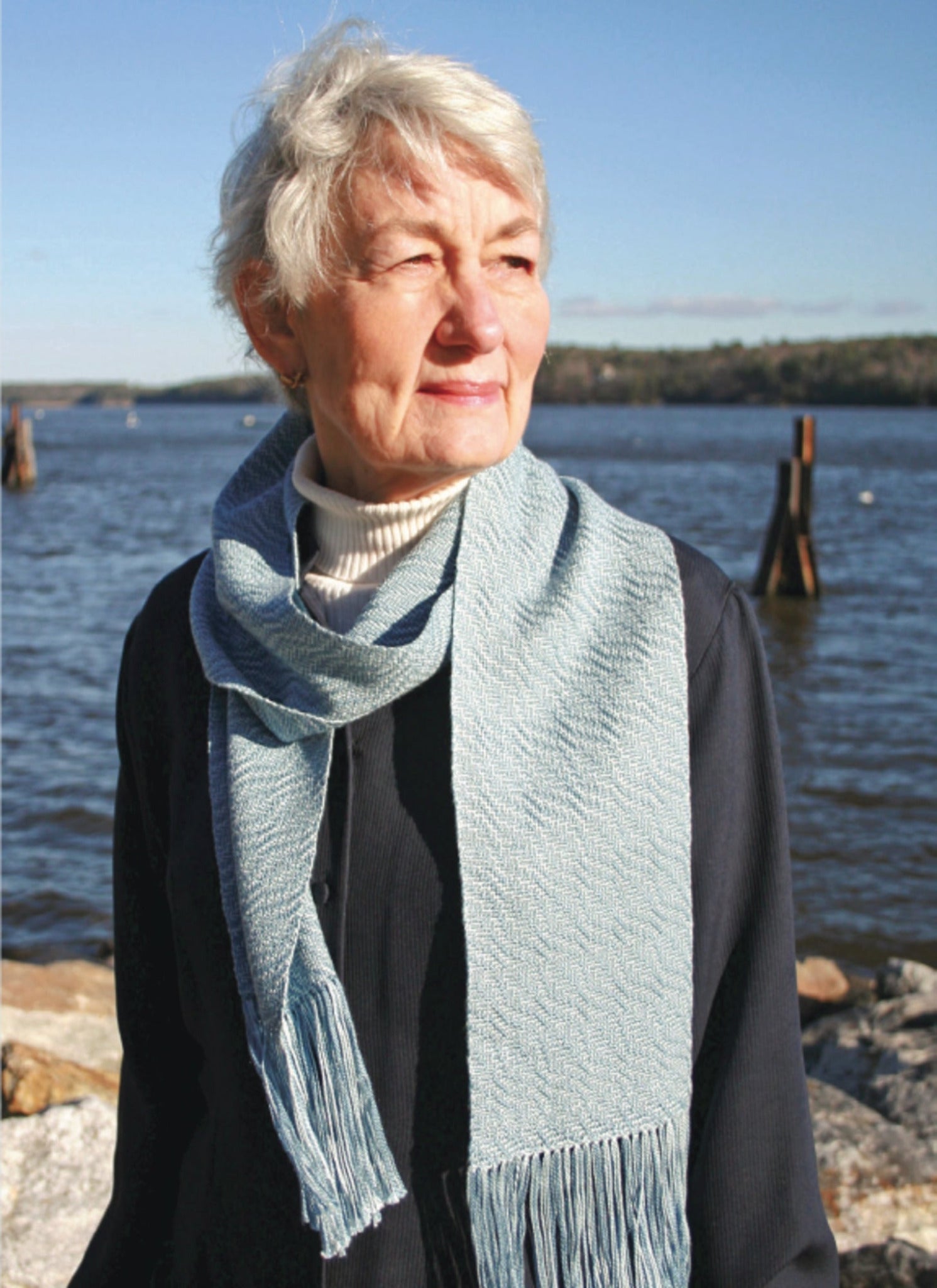 A woman with short gray hair and wearing a Snow Crystal Shadow Weave Scarf by Halcyon Yarn stands outdoors by the water. She is dressed in a dark jacket and light-colored turtleneck, looking off into the distance. The background features the shoreline and clear sky, reflecting a calm day.