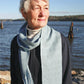 An older woman with short white hair stands near a body of water, looking to the right. She wears a dark jacket, a white turtleneck sweater, and a light blue Snow Crystal Shadow Weave Scarf from Halcyon Yarn. The background features calm water, distant land, and a clear blue sky.